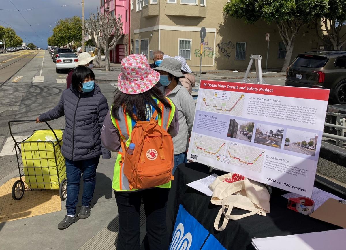 Two women talk to an SFMTA staff member about M Ocean View plans at a transit stop pop up.