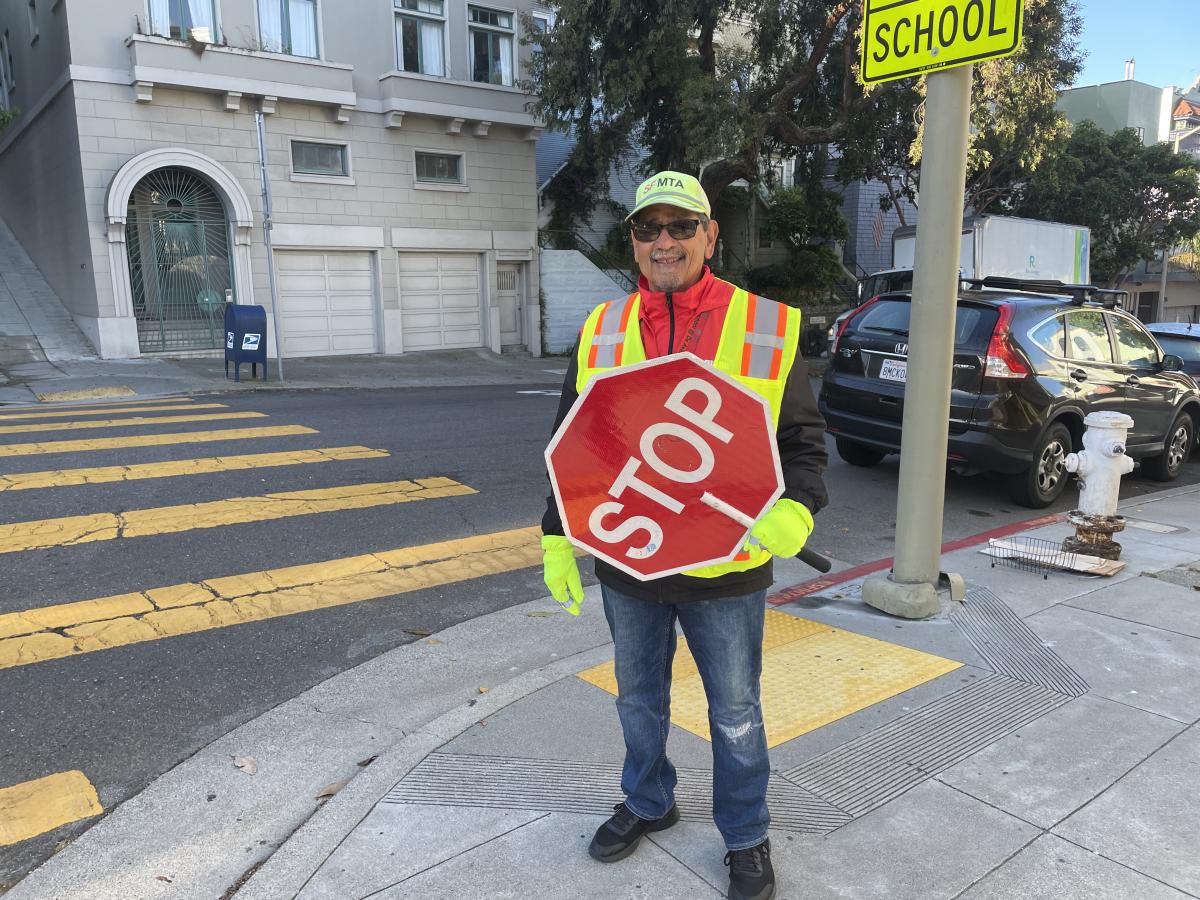 A man at a street corner wearing a safety vest and holding a stop sign.