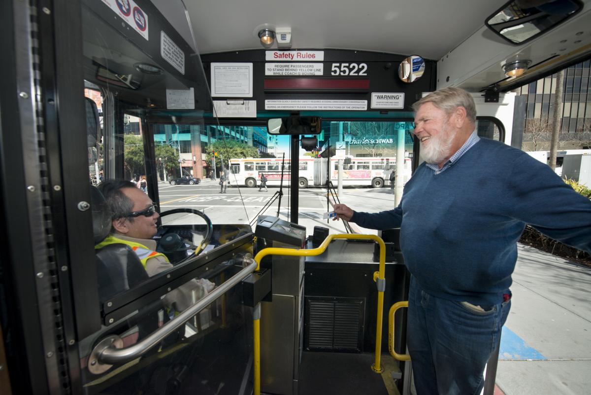 A man with a beard, sweater and jeans is smiling at the front of a bus to an operator who is wearing sunglasses seated in their seat.