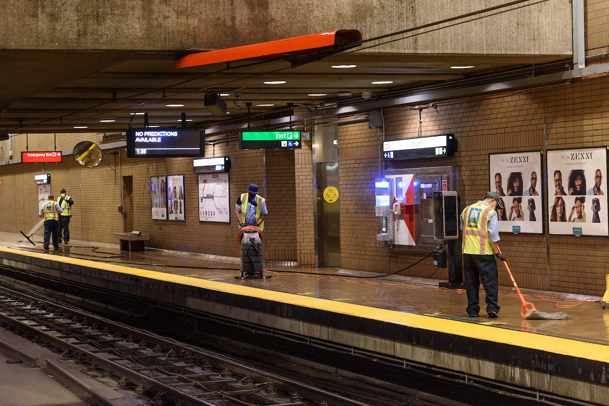 Several crew seen cleaning the ground in a station with a variety of posters and lights on the wall. 