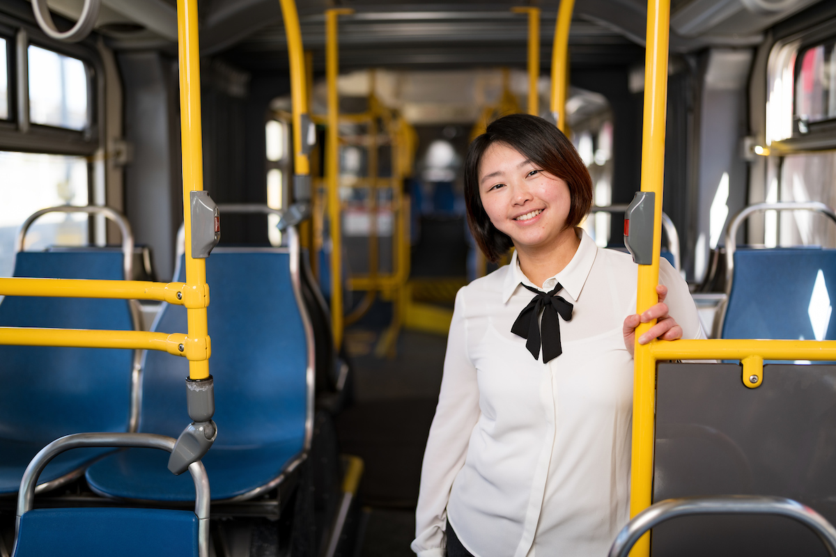 Becky smiles from inside a bus as she holds onto a pole. We see blue chairs and yellow poles near her.