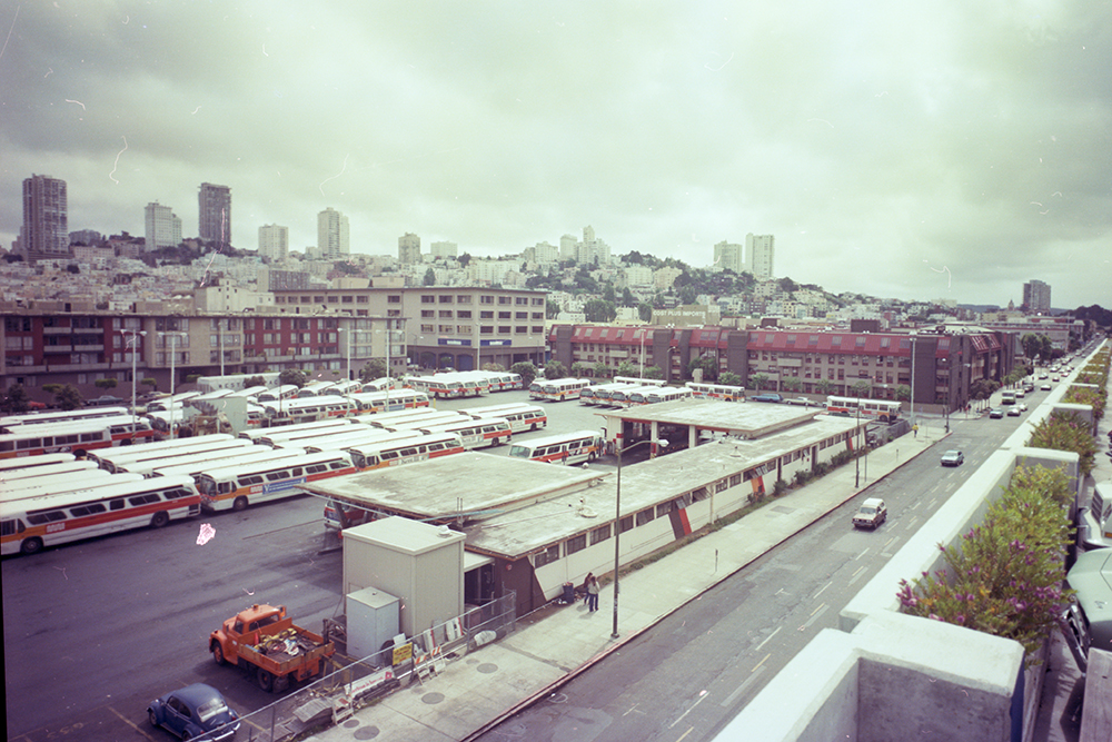 Shot of Kirkland Yard full of buses in the 1980's. Cars pass on the adjacent street. We see Nob Hill apartments in the background.