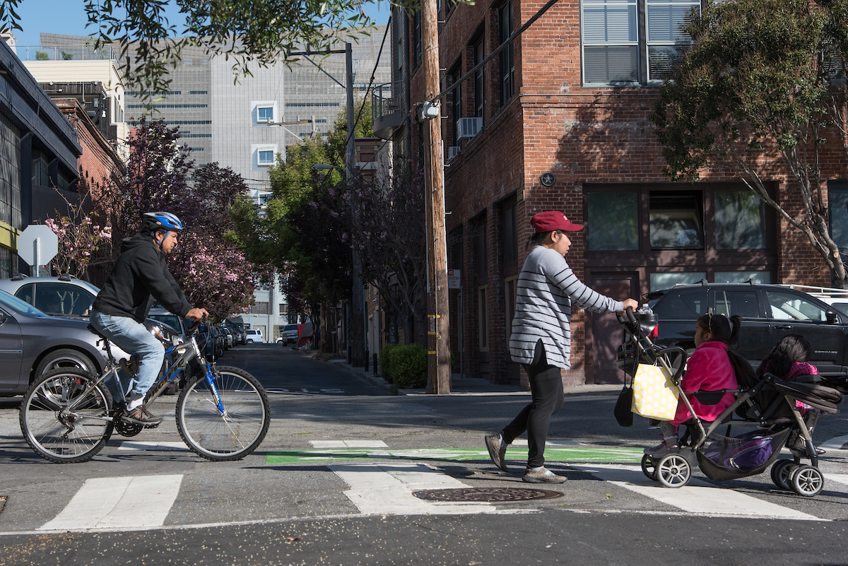 A person biking and a woman pushing a young child in a stroller cross Folsom Street. Cars are parked in the background.