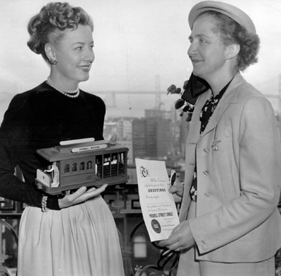 Actress Irene Dunne and activist Friedel Klussman smile with the Bay Bridge behind them; Irene holds a miniature cable car and Friedel holds a flyer