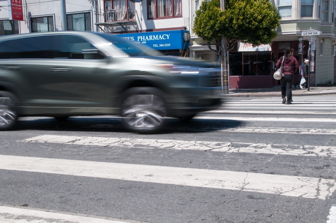 A grey car appears blurry as it speeds through a cross walk with a person crossing to the other side in the background.
