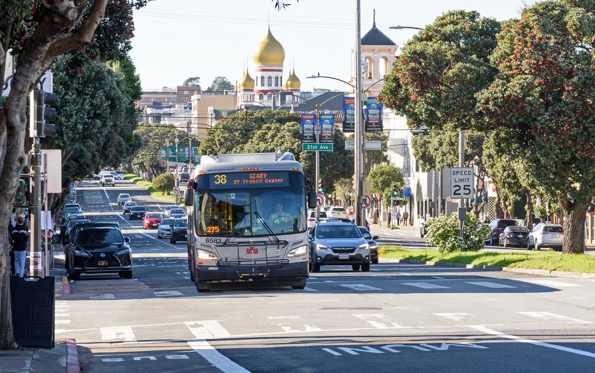 A 38 Geary bus heads east on Geary Boulevard, passing 21st Ave. in a designated transit lane. Cars follow in adjacent lanes.