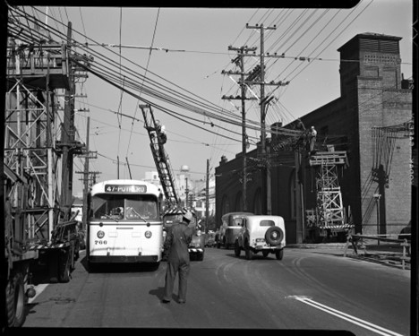 A black and white image of a bus with people working on overhead wires, one of them climbing a ladder