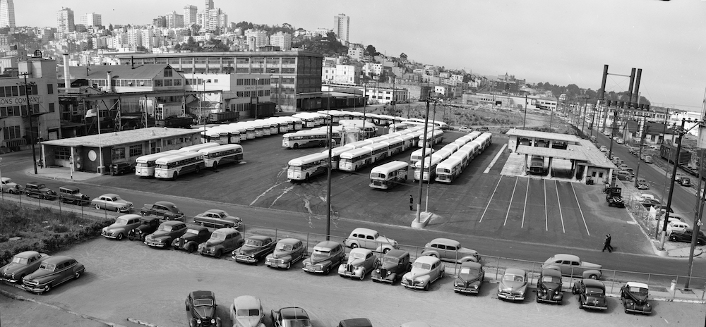 Black and white shot of Kirland Yard. Buses fill the yard and classic cars are parked beside it. Nob Hill apartments are in the background.