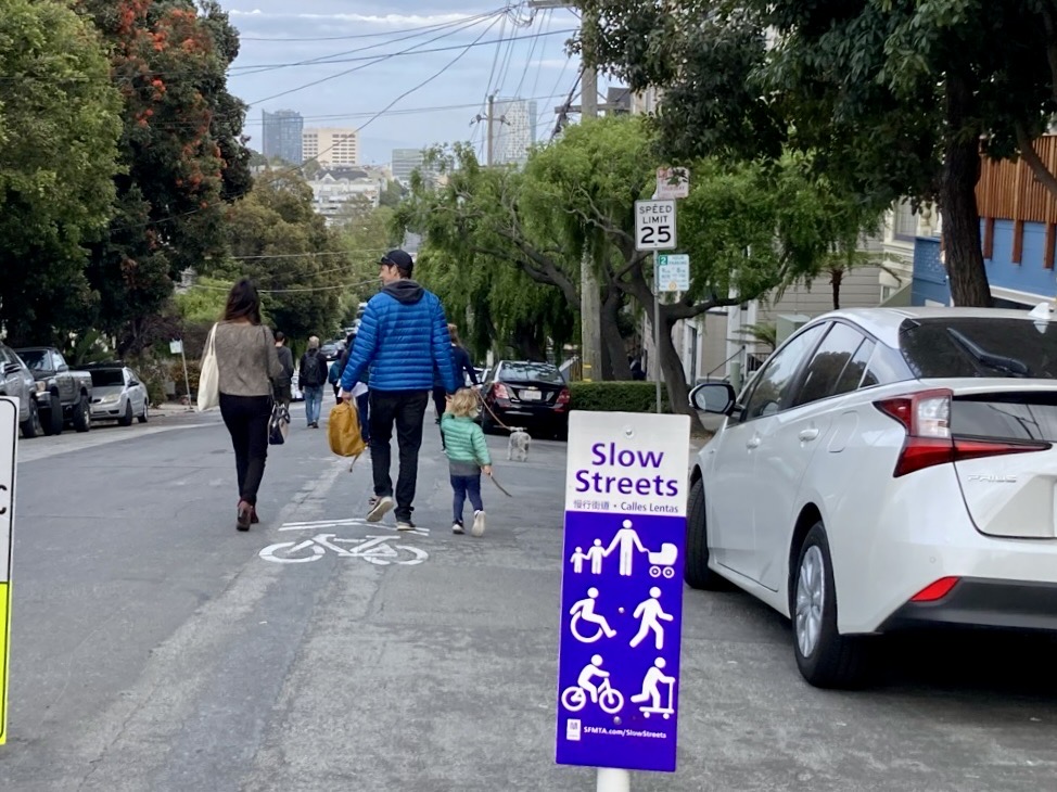 Image of a father and daughter holding hands while talking with a woman and walking in the Page Street roadway eastbound towards Divisadero Street with the San Francisco skyline in the distance