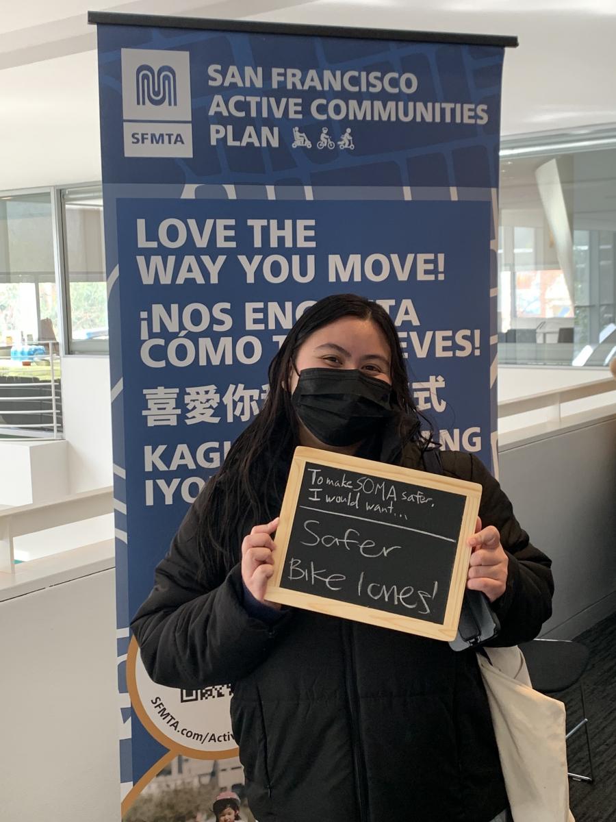 A woman holding a sign saying "To make SOMA safer, I would want ... safer bike lanes!"