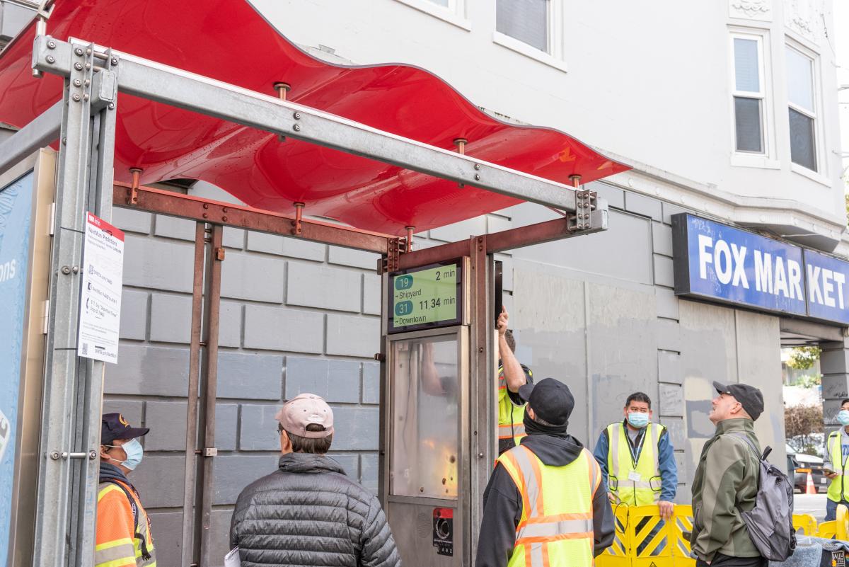 A picture containing contractors speaking with a Muni Rider as customer information displays are being installed in a transit shelter. 