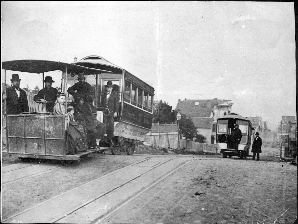 Black and white photo of two cable cars with people riding on the back and looking at the camera.
