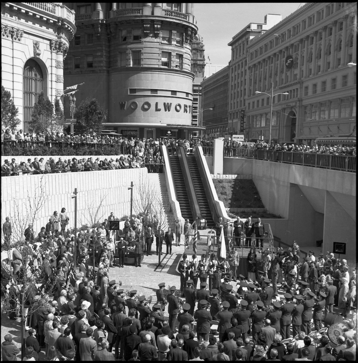 Black and white photo of a ceremony at the bottom of an escalator with crowds watching a speaker.