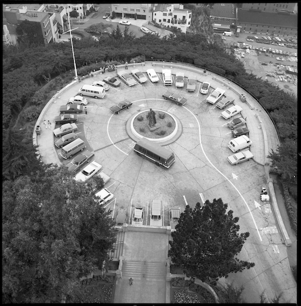 A black and white overhead photo of a circular parking lot full of cars and buses and surrounded by trees.