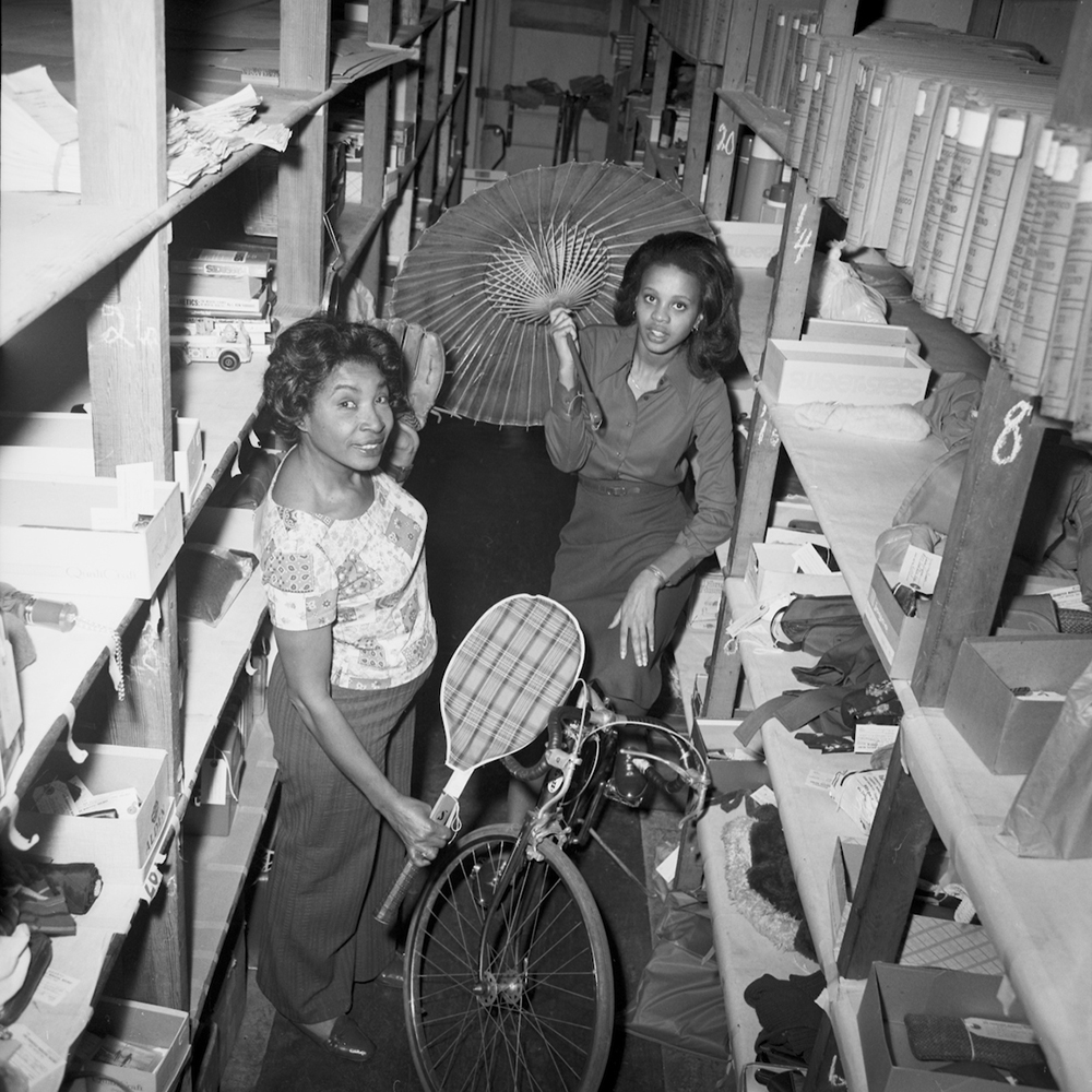 Employees standing in between shelving holding various items such as a umbrella, tennis racket and bicycles. The shelving is filled with additional various items. 