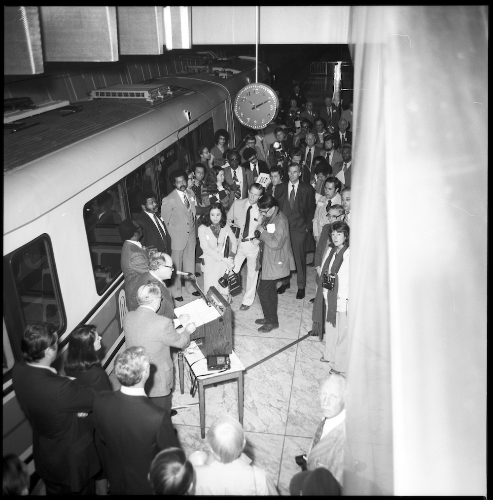 black adnw hite photo looking down to platform at Embarcadero Station to crowd of people next to LRV and speaking podium