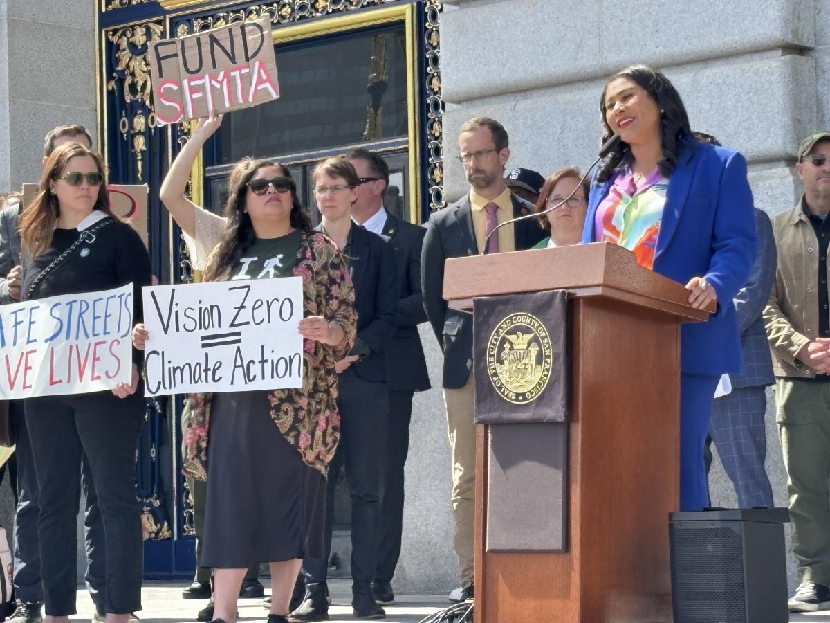 Mayor Breed speaks at a podium in front of city hall. People stand beside her holding signs that say "Fund SFMTA" and "Vision Zero = Climate Action."