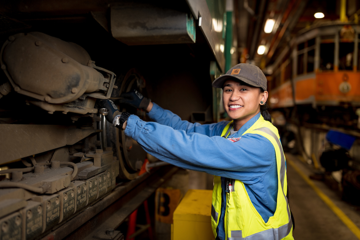 Jeena Villamor adjusts track brakes for a historic streetcar. She wears a yellow safety vest and blue uniform and makes a repair.