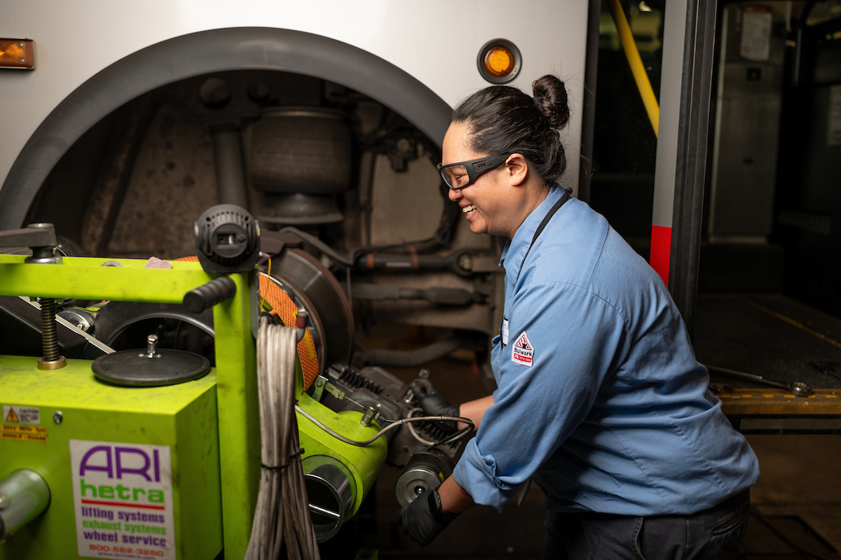 Jenny measures break rotors for a vehicle. We see her smiling and wearing a blue uniform as she works on a green machine.