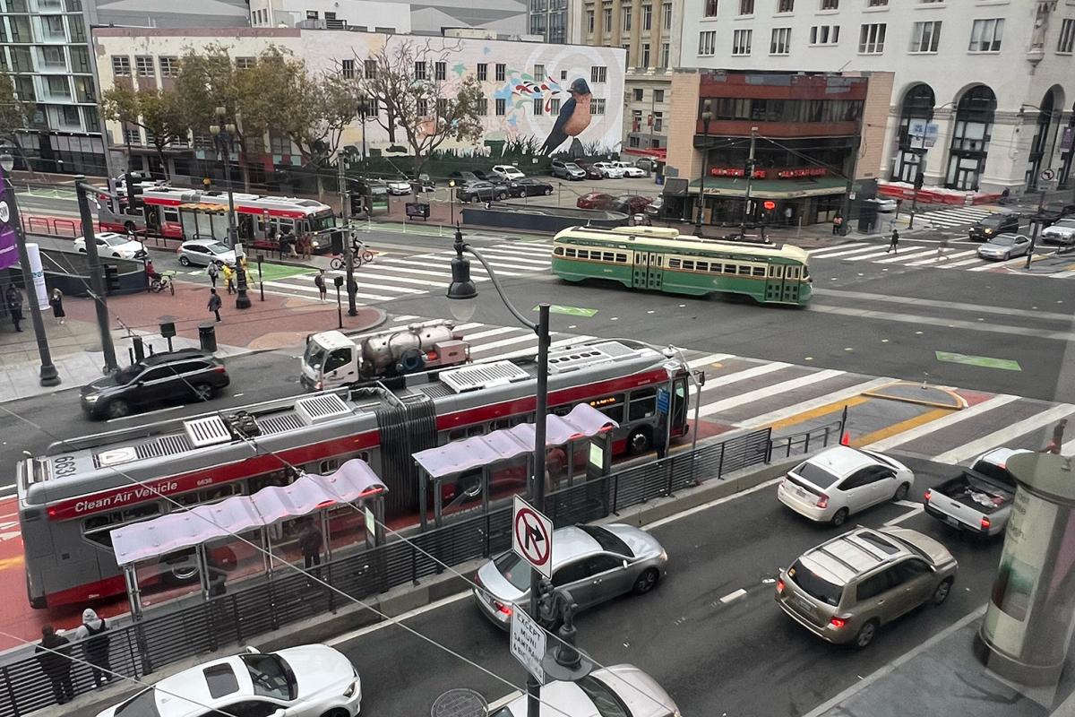 Image of a busy intersection corner possibly during commute showing multiple cars and buses, pedestrians and a trolley 