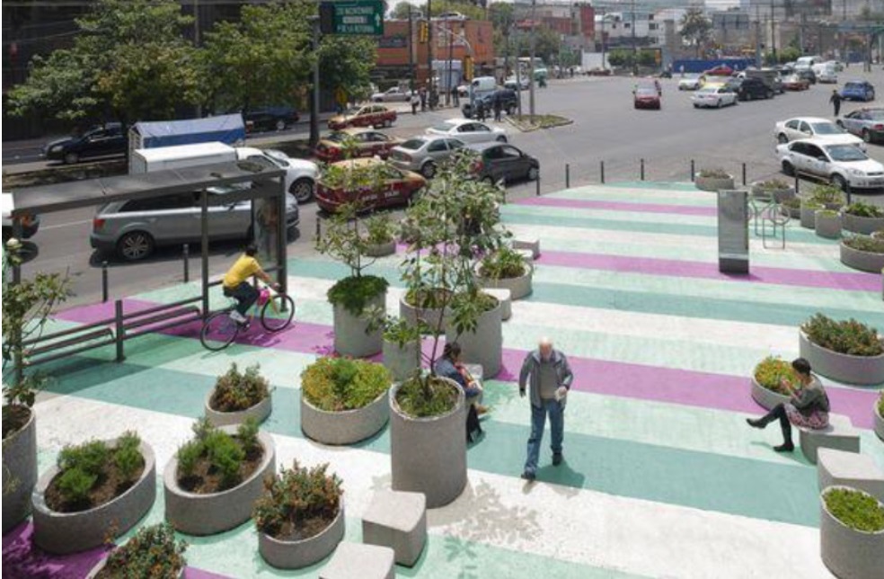 Street with some space reserved for pedestrians defined by bollards on the outside and a painted mural and planters holding trees and other plants.