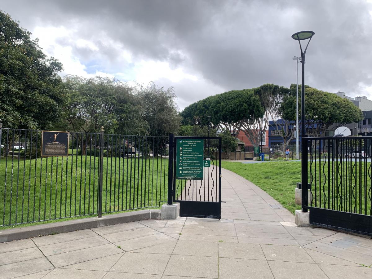 Image shows a concrete pathway and the open gate of Victoria Manalo Draves Park, which lead to gentle grassy hills and a basketball court