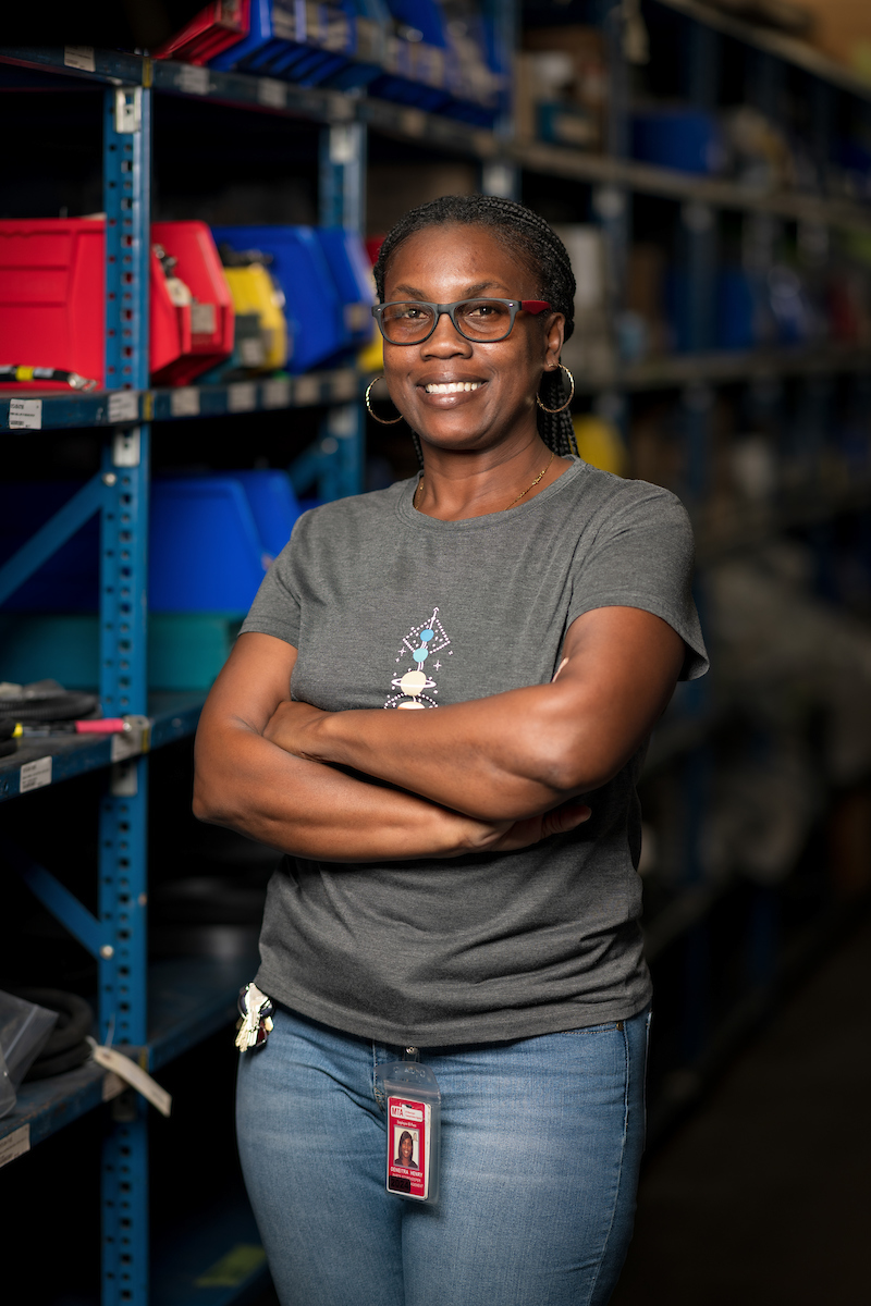 Deneitra Henry smiles from inside a parts storeroom. We see shelves of parts behind her.