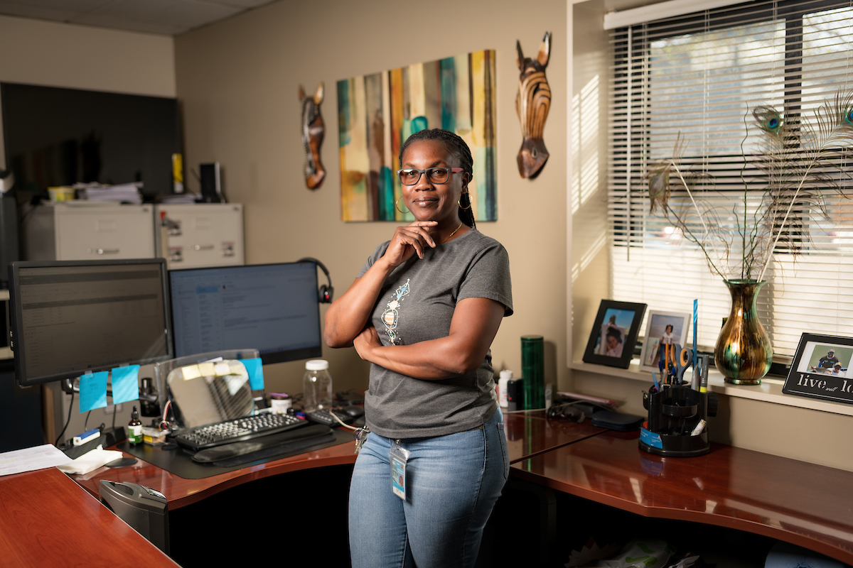 Deneitra Henry smiles from inside her office. We see her desk, computer, photos and other items in the background.