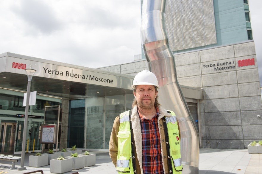 Man wearing hardhat and vest outdoors in front of transit station.