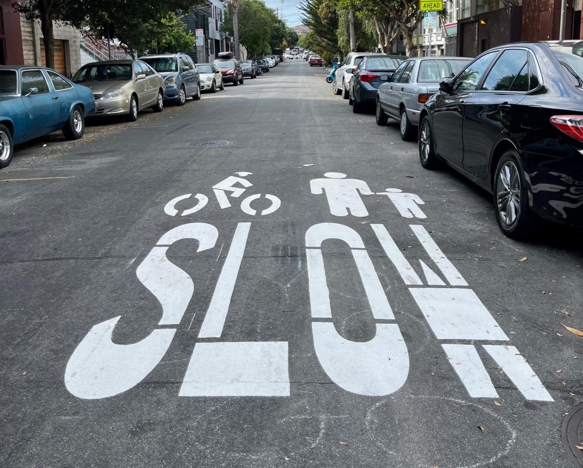 Image of roadway with paint sign that says "Slow" and includes an image of a cyclist and an adult with a child.