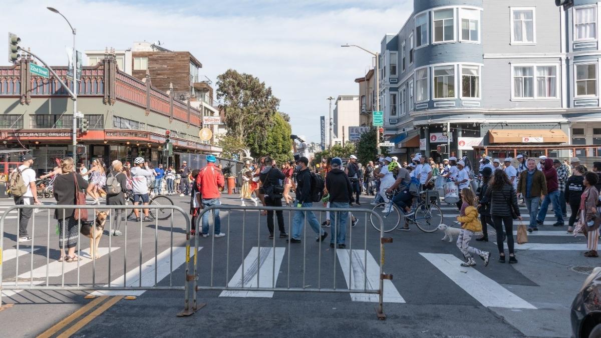 A crowd of people at a street fair with blockades in the foreground and businesses in the background.