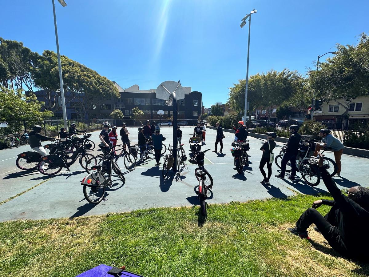 Photo of a group of Western Addition / Fillmore residents and SFMTA staff with bicycles having a conversation at a basketball court. 