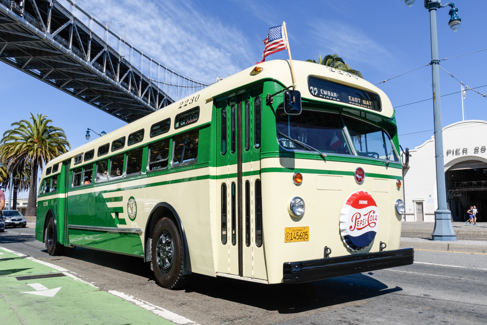 A vintage Muni bus that's green and cream in color passing under a bridge 