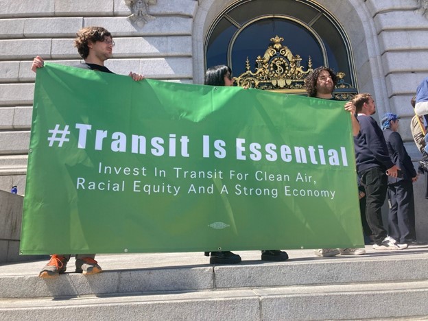 A group of people hold a banner that says Transit Is Essential, Invest in Transit For Clean Air, Racial Equity and a Strong Economy