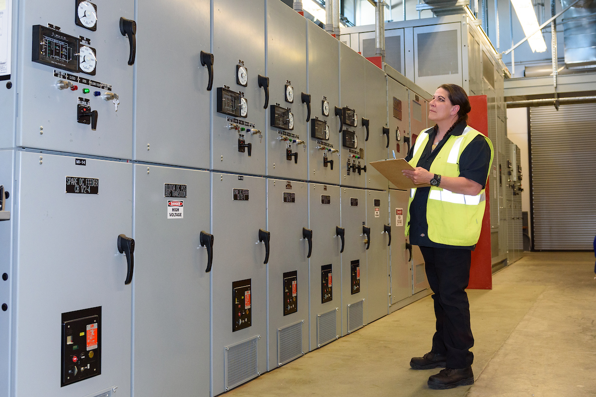 Ess stands in a substation. We see her in the background holding a clipboard as she observes machines.