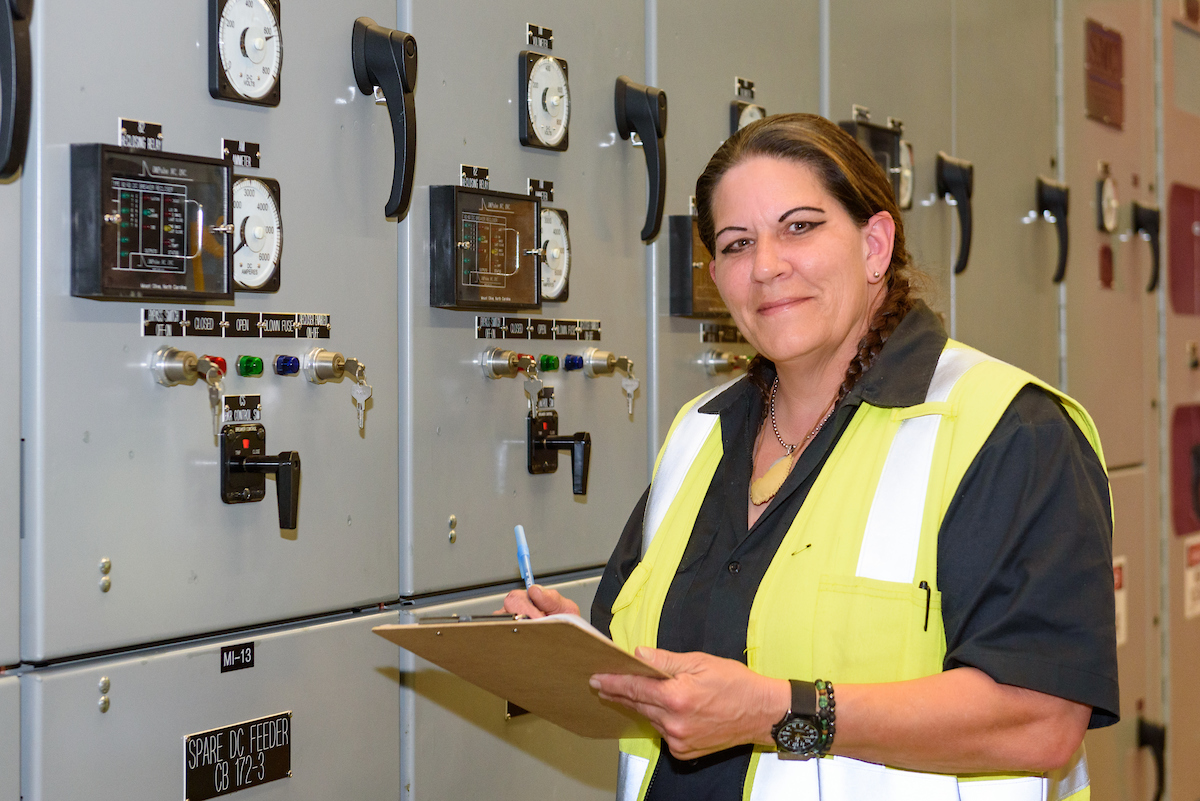 Ess records data from a substation. She wears a yellow vest and holds a clipboard as she smiles at the camera.