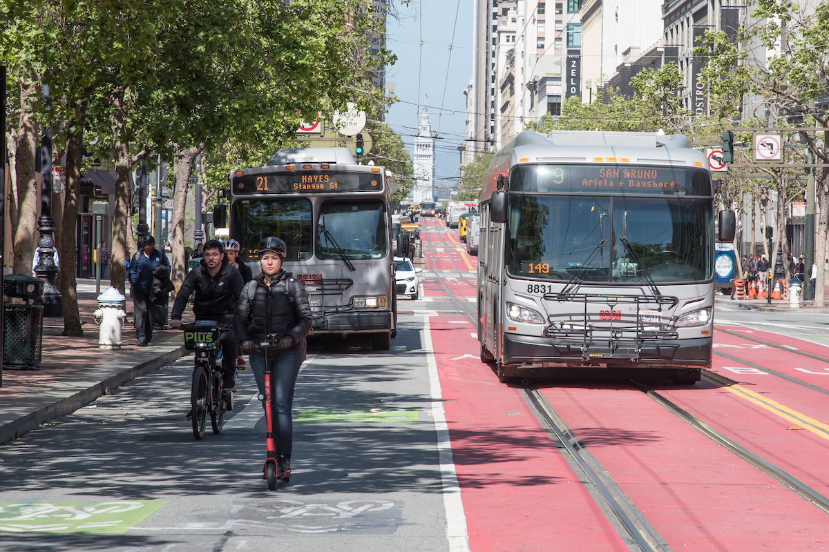 Two buses, a bicyclist, a pedestrian and person riding a scooter all on Market Street.