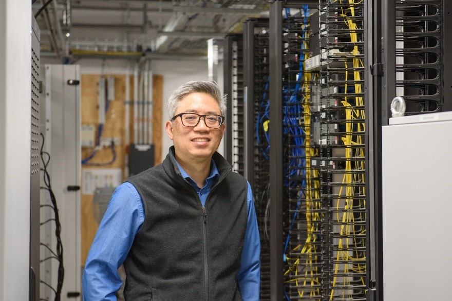 Man in blue shirt and grey vest standing in server room.