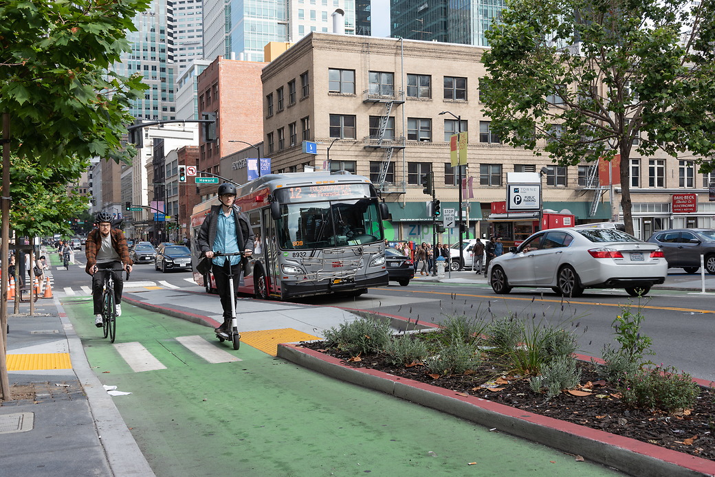 A person rides a scooter in the protected 2nd Street bike lane alongside a cyclist. A Muni bus is visible in the background.