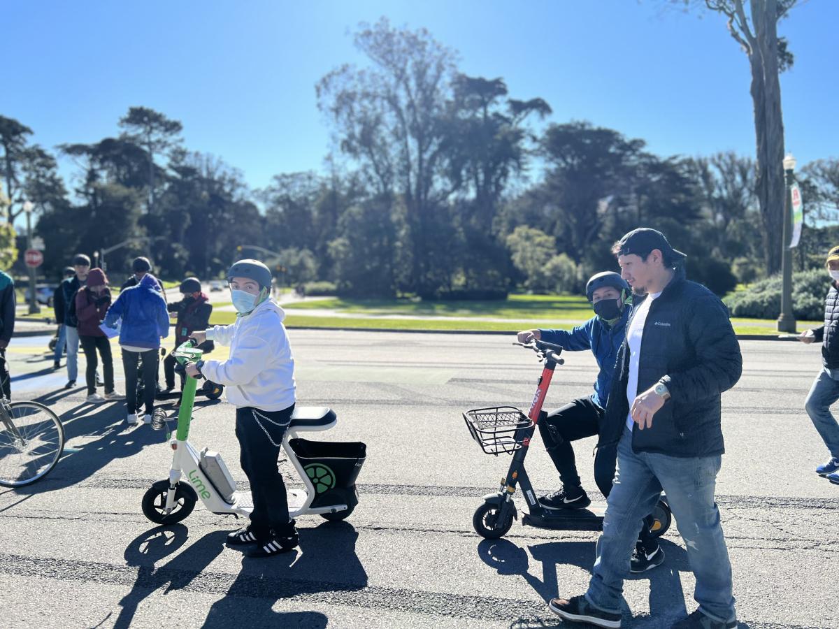 Two students ride adaptive scooters in Golden Gate Park as a man walks alongside them.