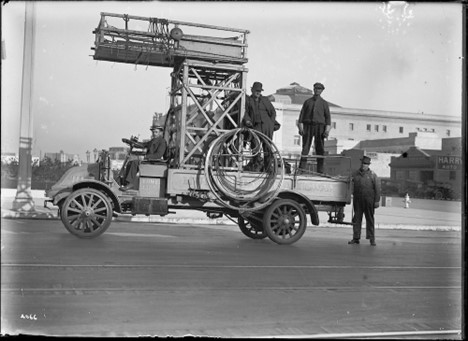 A black and white image of an old railway tower truck with a man in the driver seat, two men standing on the truck bed, and one man standing behind the truck