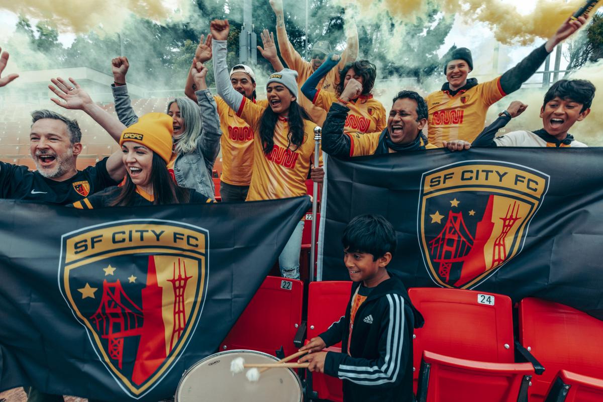 Fans of the San Francisco City Football Club cheer on the home team at Kezar Stadium. Fans wear yellow-orange jerseys with the Muni logo and hold banners. We see thick fog and trees behind them.