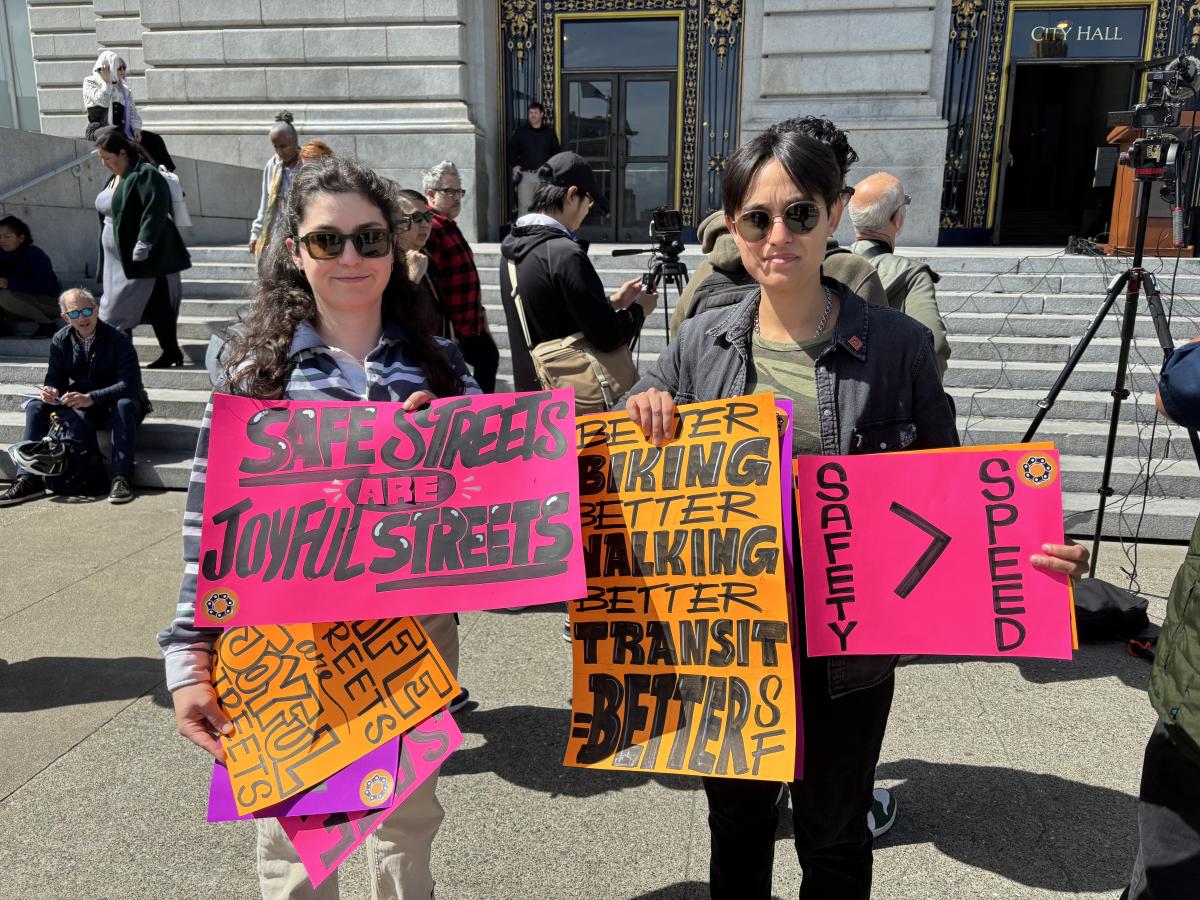 Two people stand in front of SF City Hall holding signs that say Safe Street are Joyful Streets and Safety Over Speed.