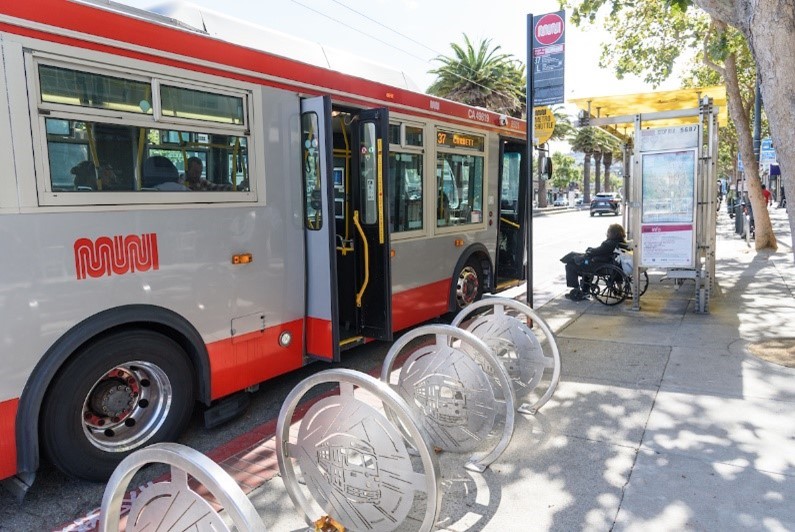 Muni bus with doors open waiting at a bus stop and picking up a person in a wheel chair. There is bicycle parking and an overhead shelter at the stop.