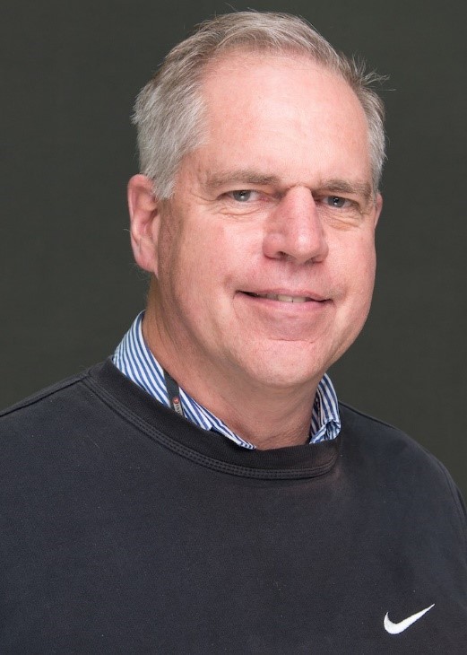 Man in front of grey background wearing blue sweatshirt and blue striped shirt.