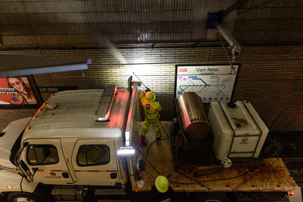 Crew member on the back of a truck spraying water along the side of the tunnel wall. There is a map on the right side and a advertisement board on the left  of a woman with sunglasses smiling.