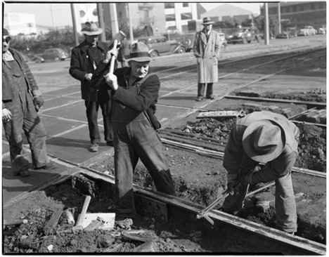 Black and white photo of two people working on tracks. One holds a rail spike while the other strikes it with a large hammer