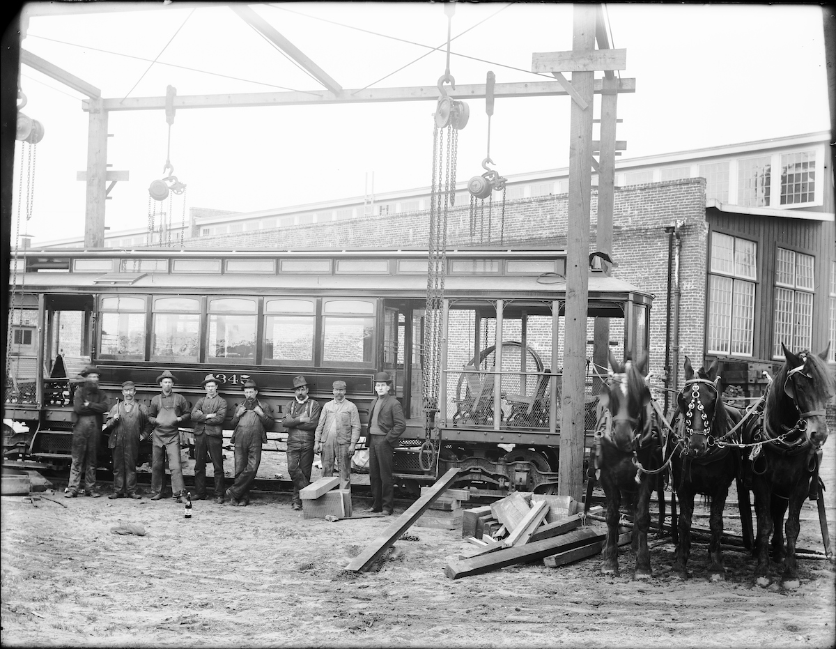 A black and white photo of an early 20th Century streetcar surrounded by a work crew and horses.