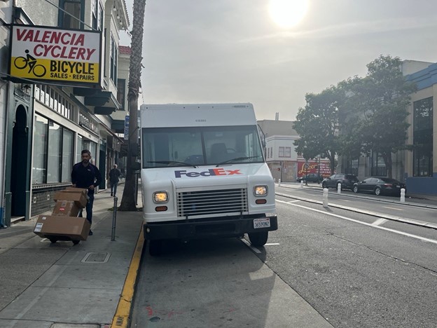 A FedEx delivery truck pulled to the side of the road with the driver moving boxes on a dolly loader.