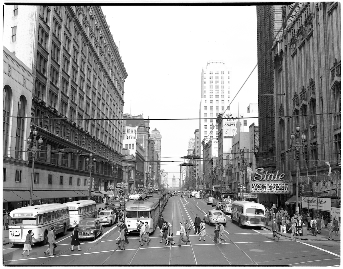 A black and white vintage photo of a busy street with buses and people crossing the street.